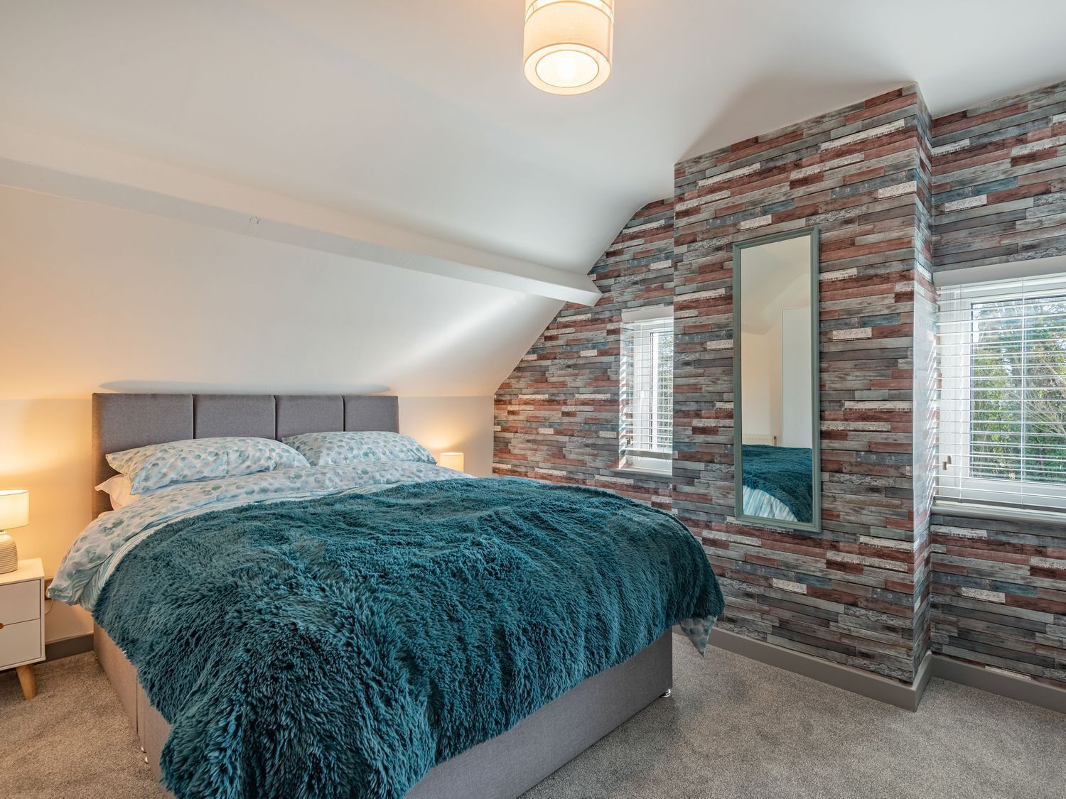 A bedroom with a bed, a nightstand, a wall-mounted mirror, and a window with blinds.