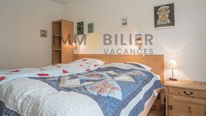 A bedroom with a bed, a nightstand, a lamp, and a wardrobe. There are wall decorations and a sign with the text "IMMOBILIER VACANCES RÉSEAU MANTIS". - Très agréable 2 pièces contemporain, idéal pour 2 à 4 personnes, situé dans une petite résidence de (St-Luc)
