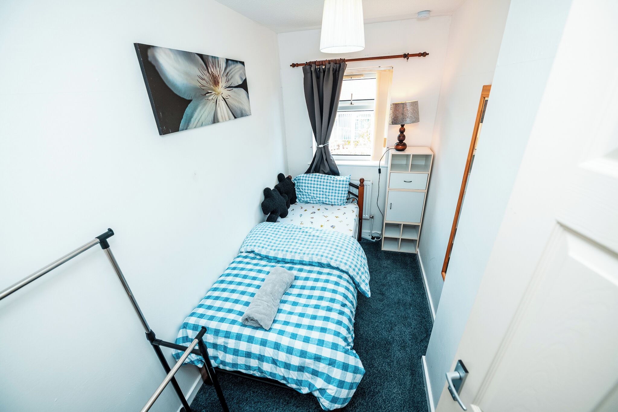 Una habitación pequeña y estrecha con una cama, un escritorio y una silla. Hay una ventana con cortinas, una lámpara y un cuadro en la pared.