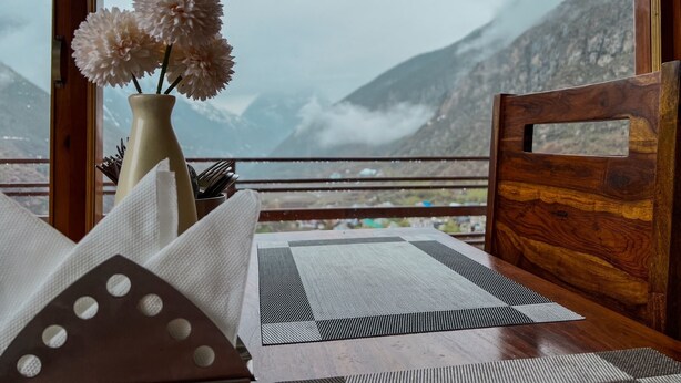 Une table en bois avec un set de table, un vase rempli de fleurs et une vue sur les montagnes par la fenêtre.