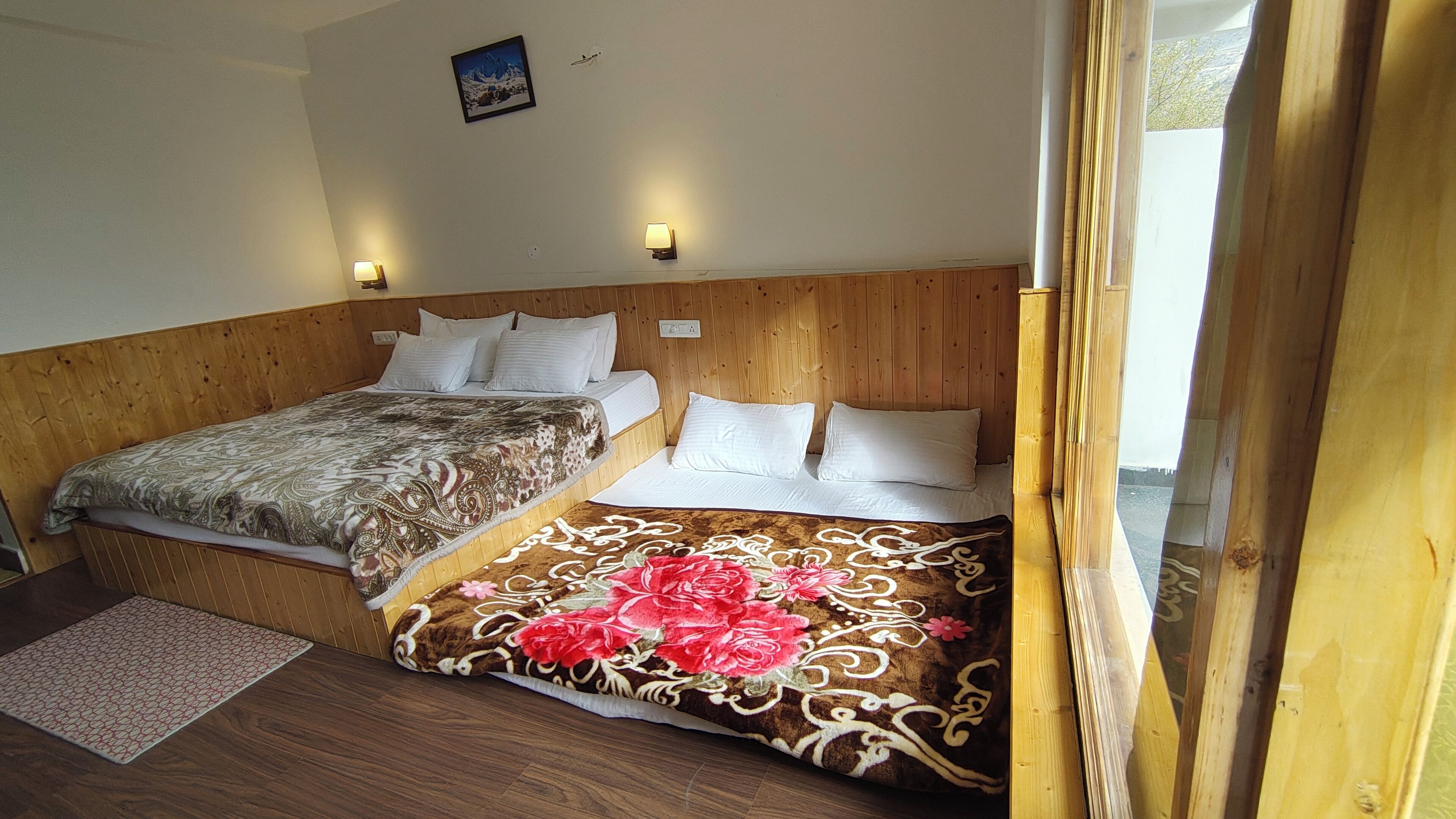 A bedroom with two beds, wooden headboard, bedside lamps, a small rug, and a framed picture on the wall.