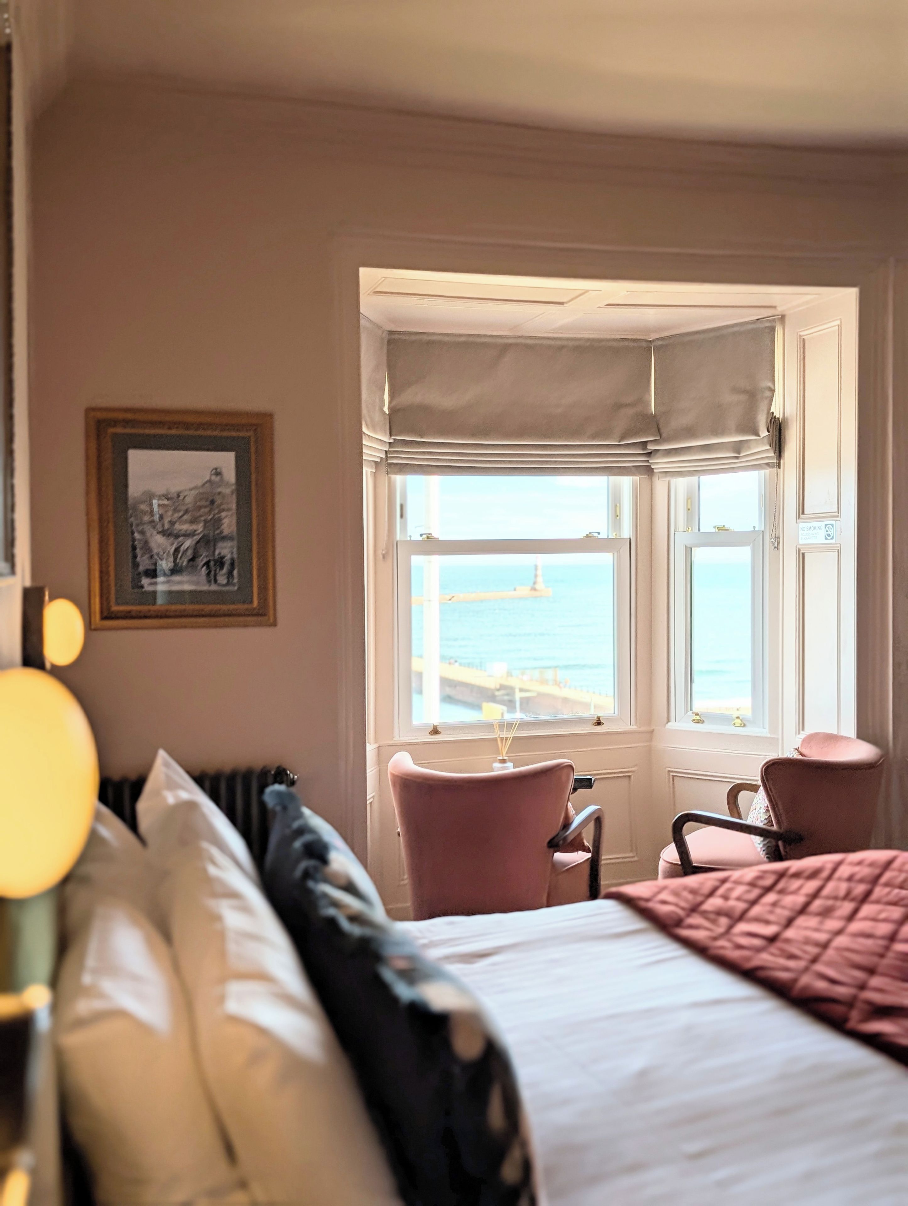 A hotel room with a bed, a chair, a window with a view of the sea, and a framed picture on the wall.