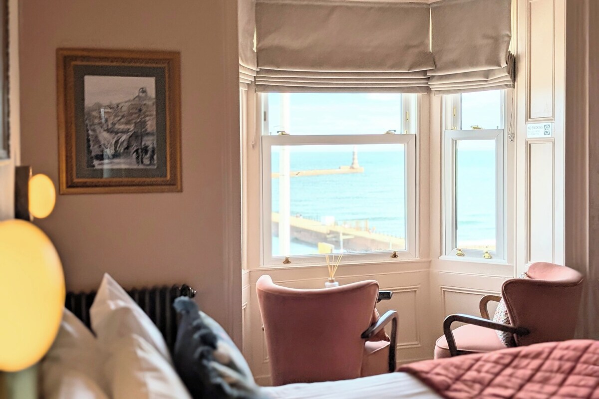 A hotel room with a bed, a chair, a window with a view of the sea, and a framed picture on the wall.