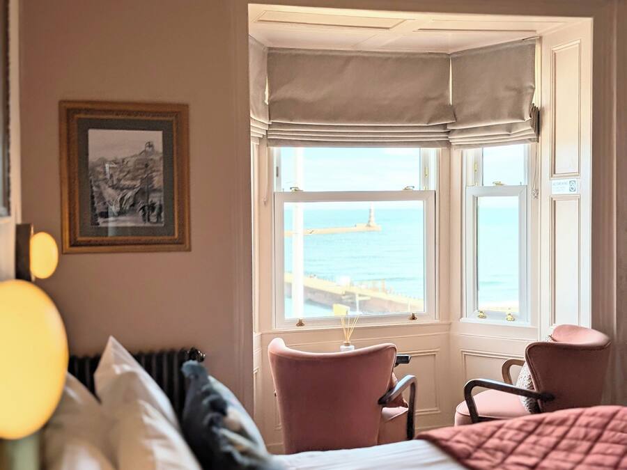 A hotel room with a bed, a chair, a window with a view of the sea, and a framed picture on the wall.