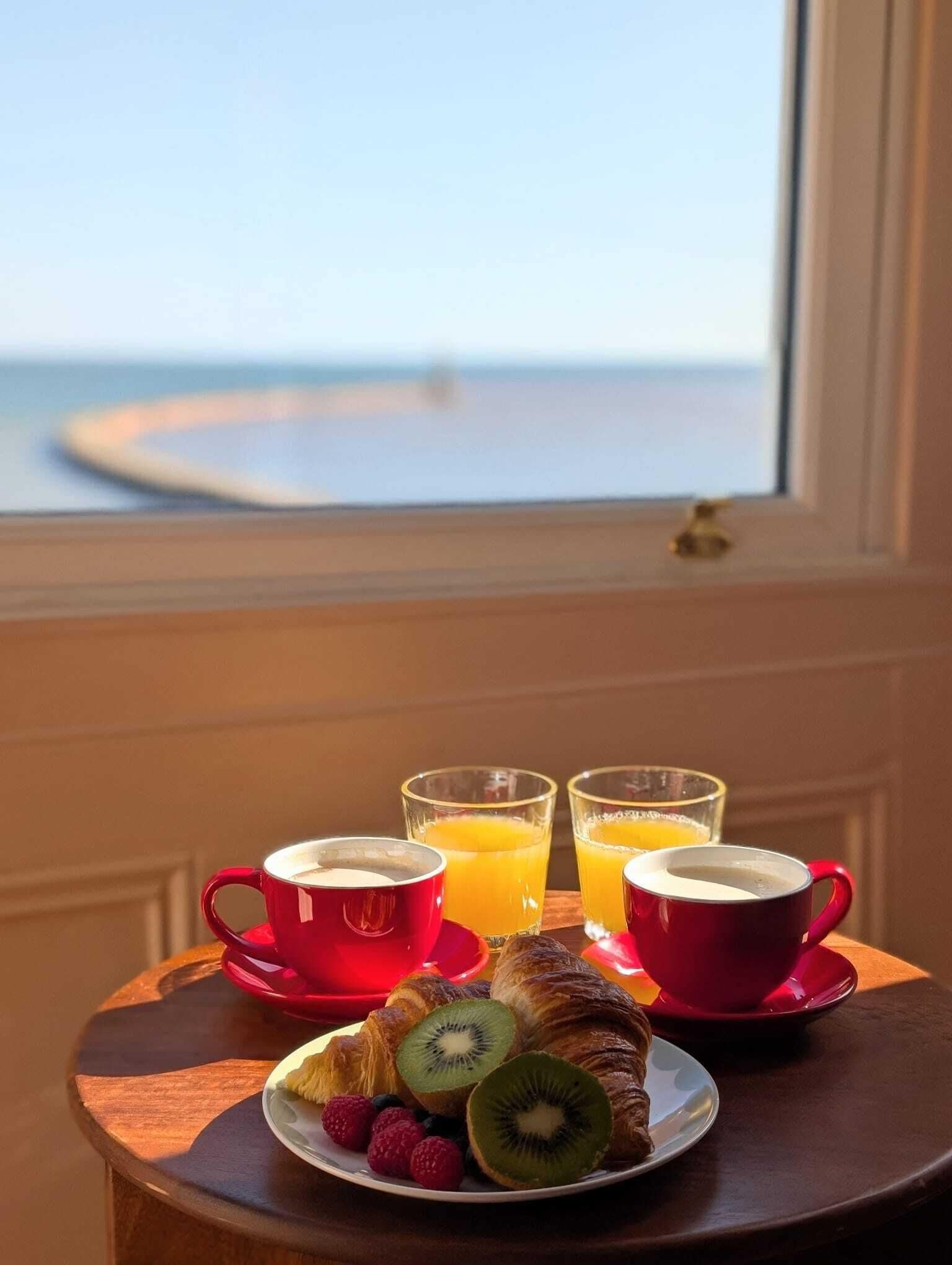 Une table de petit-déjeuner avec des croissants, des fruits et du jus d’orange, donnant sur une plage.