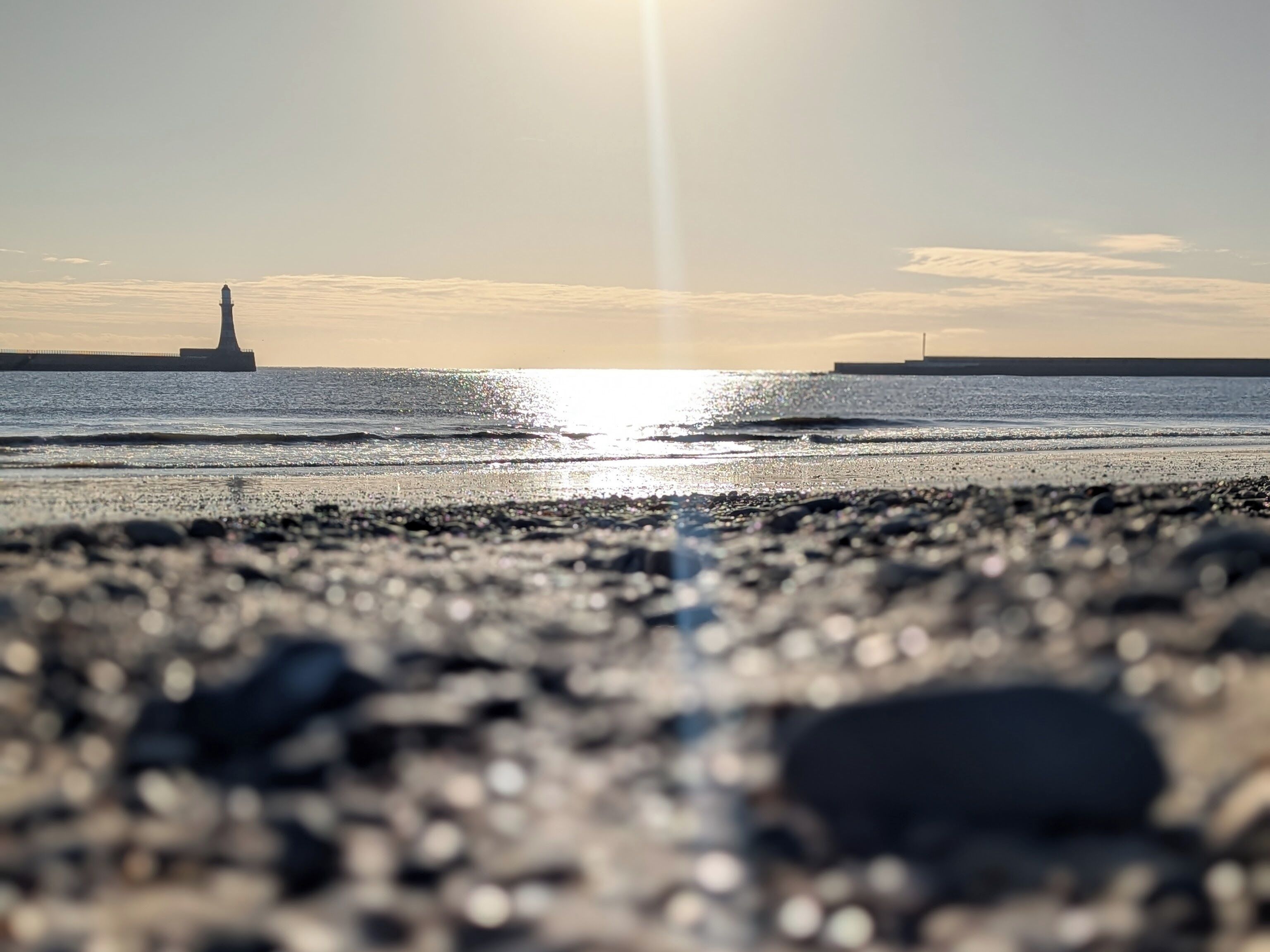 A beach with pebbles, a lighthouse in the distance, and the sun reflecting on the water.