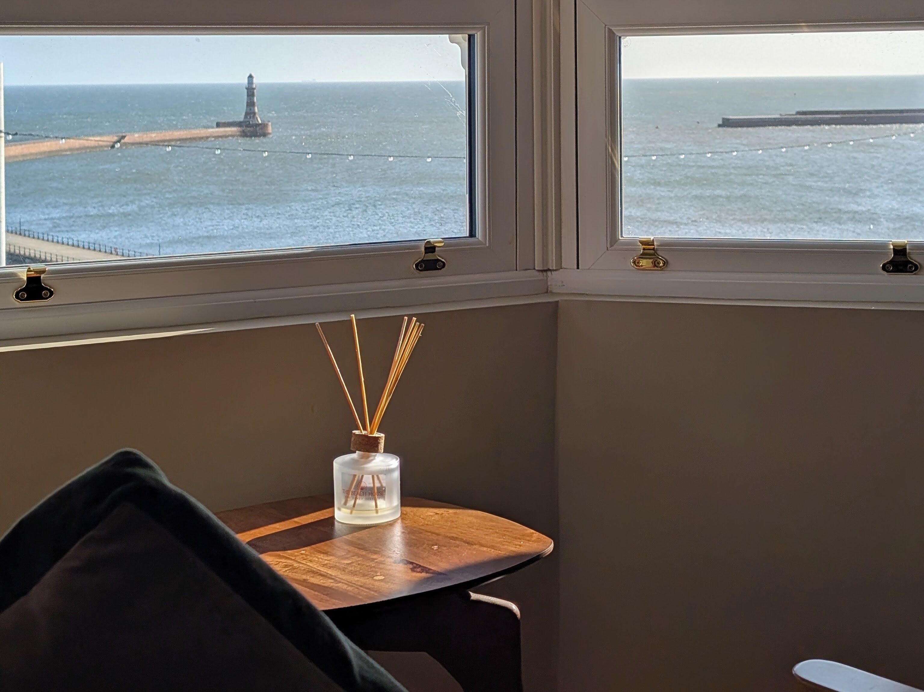 A room with a view of the sea, a lighthouse, and a table with a reed diffuser.