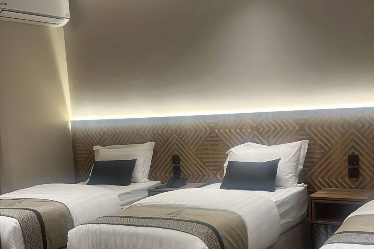 A hotel room with three beds, a patterned carpet, a ceiling light fixture, and a wall-mounted air conditioning unit.