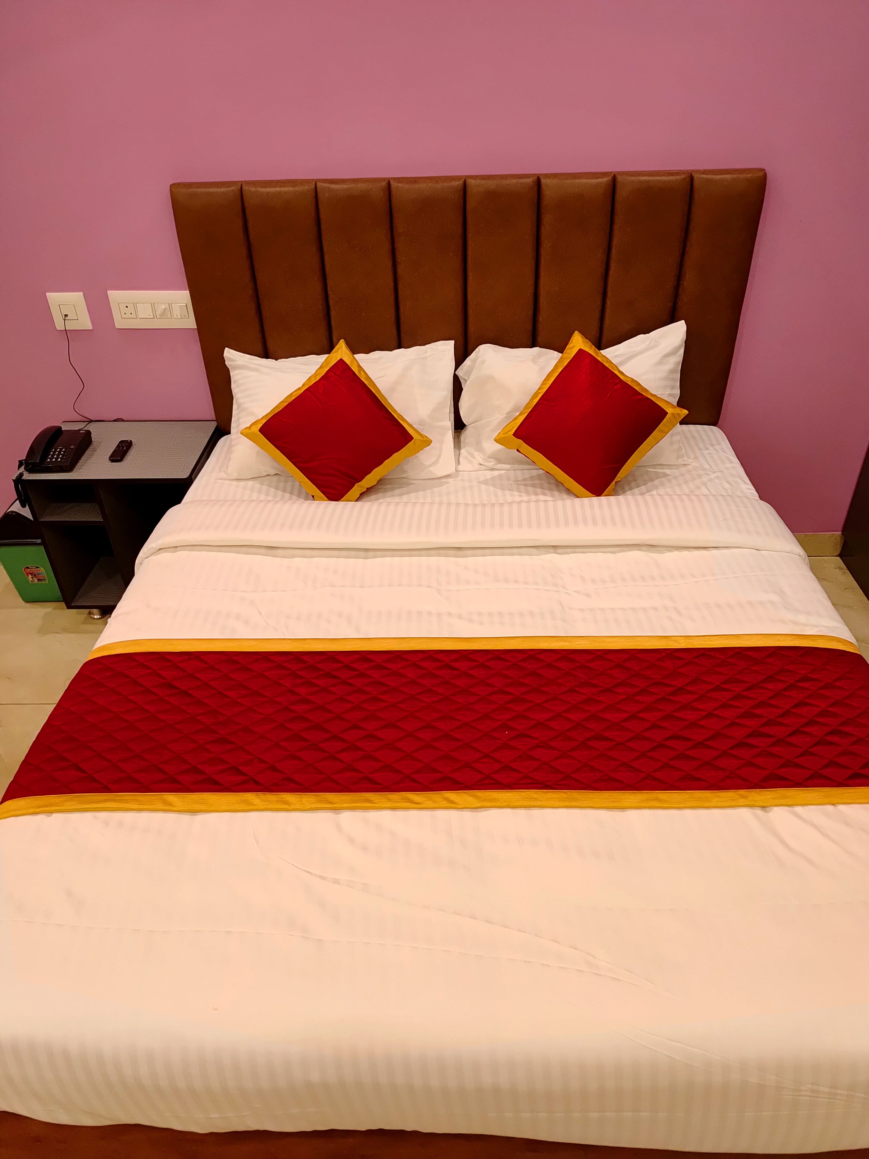 Un lit bien fait, recouvert d’une couvre-lit rouge et blanche, avec deux oreillers rouges ornés de galons dorés et une tête de lit marron recouverte de tissu.