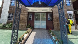 Entrada de un hotel con un toldo de vidrio azul, un cartel que dice "Welcome to [Hotel Name]" y un guardia de seguridad parado afuera.