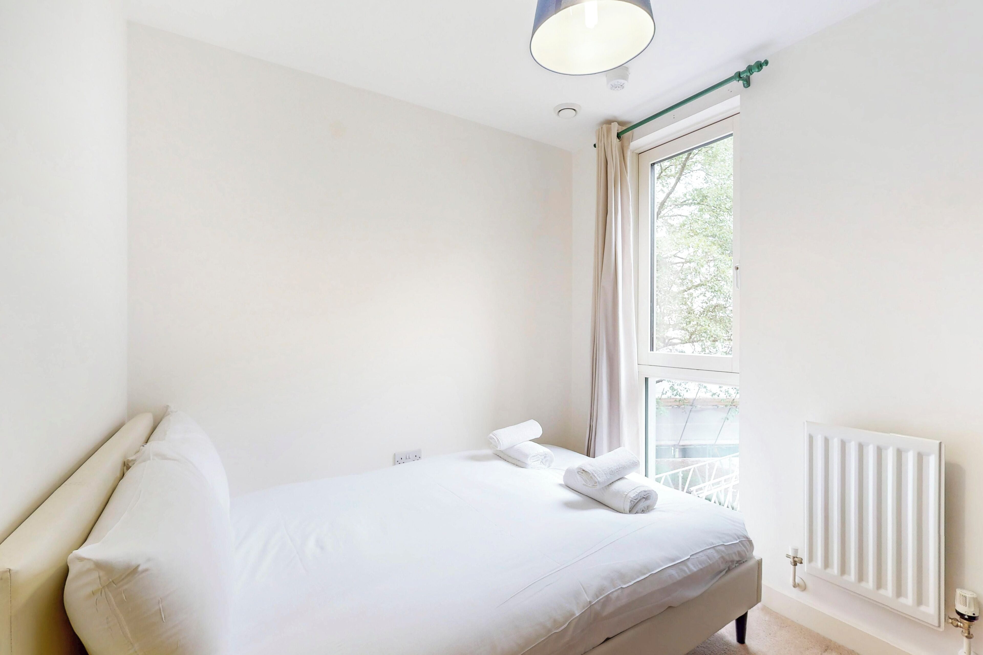Una camera da letto minimalista con un letto singolo, una finestra con tende, un termosifone e una lampada a soffitto.