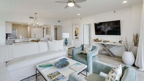 Ein modernes Wohnzimmer mit einem weißen Sofa, einem blauen Sessel und einem Couchtisch. Eine Deckenventilator, ein an der Wand montierter Fernseher und ein durch eine offene Tür einsehbarer Küchenbereich sind ebenfalls vorhanden.