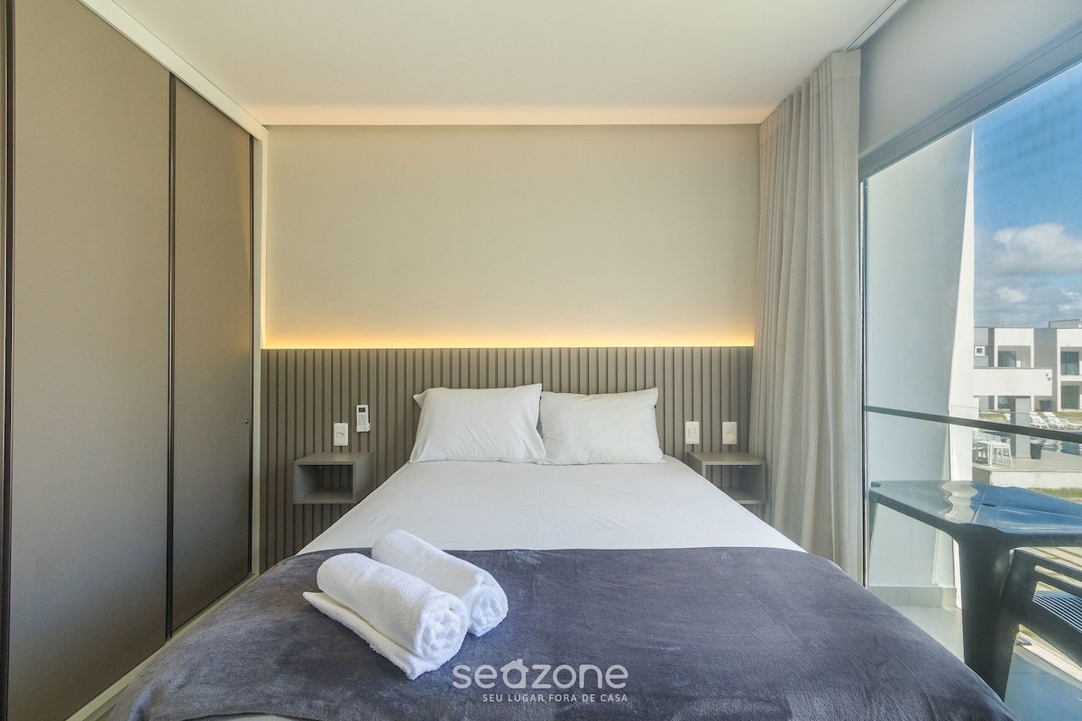 A hotel room with a large bed, a view of the city through a balcony, and a 'sedzone' sign on the bedspread.