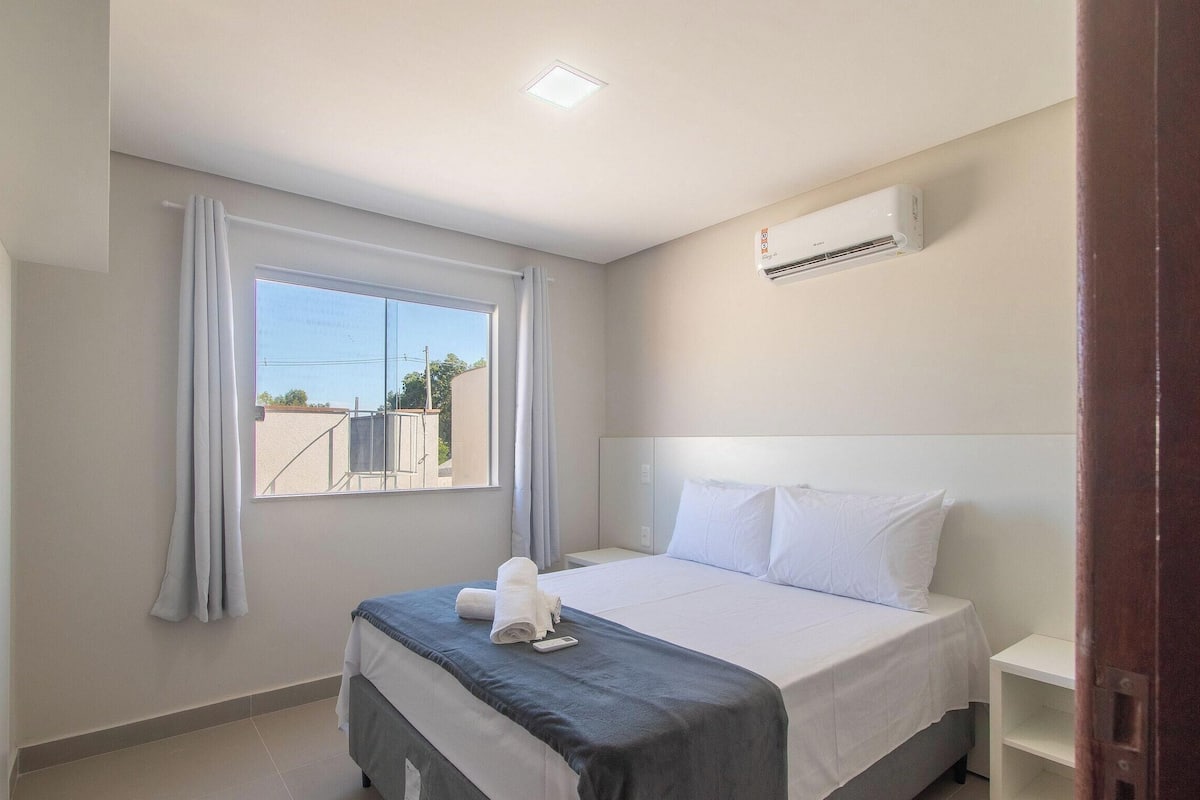 A hotel room with a bed, a window with curtains, an air conditioning unit, and a bedside table.