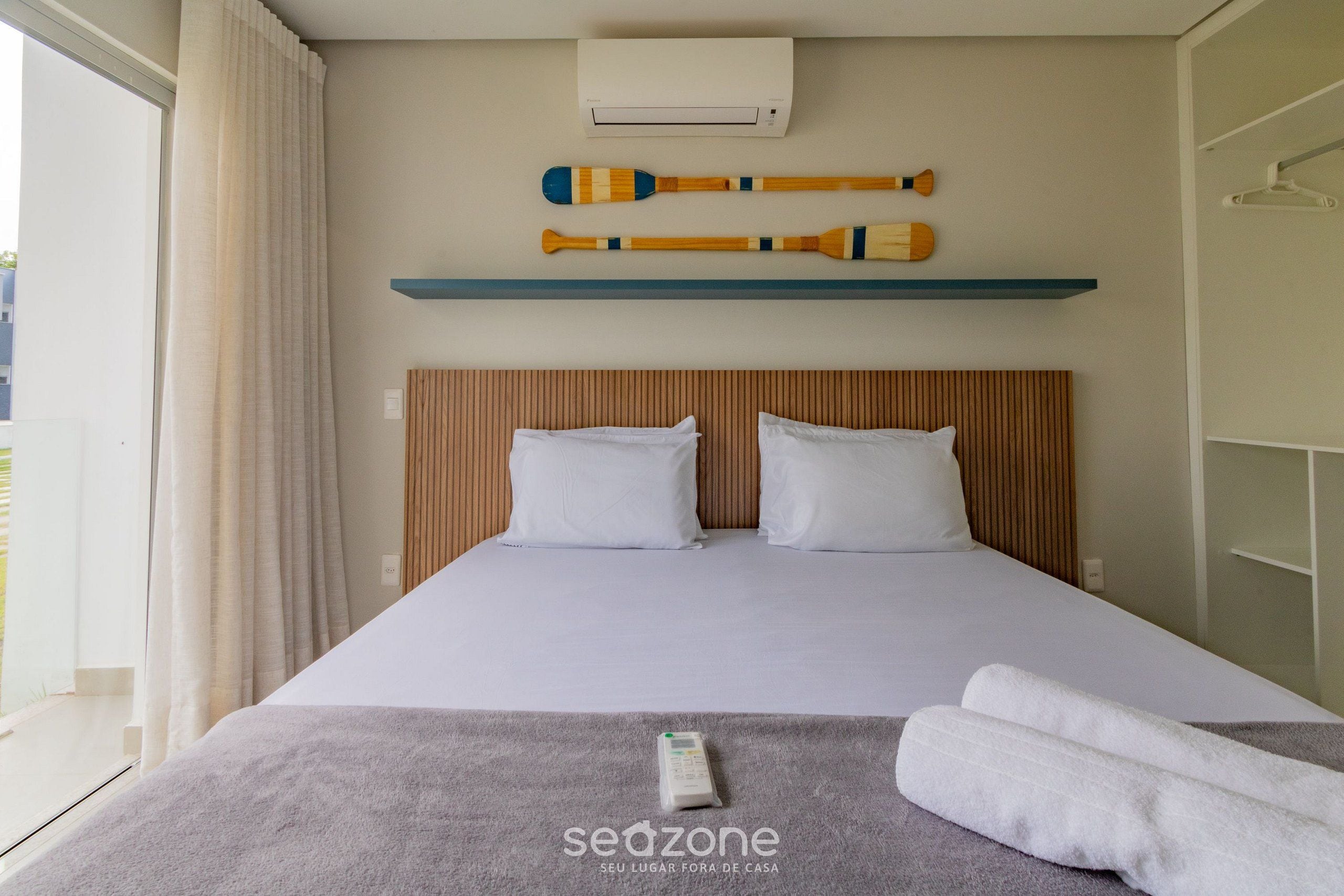 A hotel room with a bed, pillows, a remote control, towels, a shelf with batons, and an air conditioning unit.