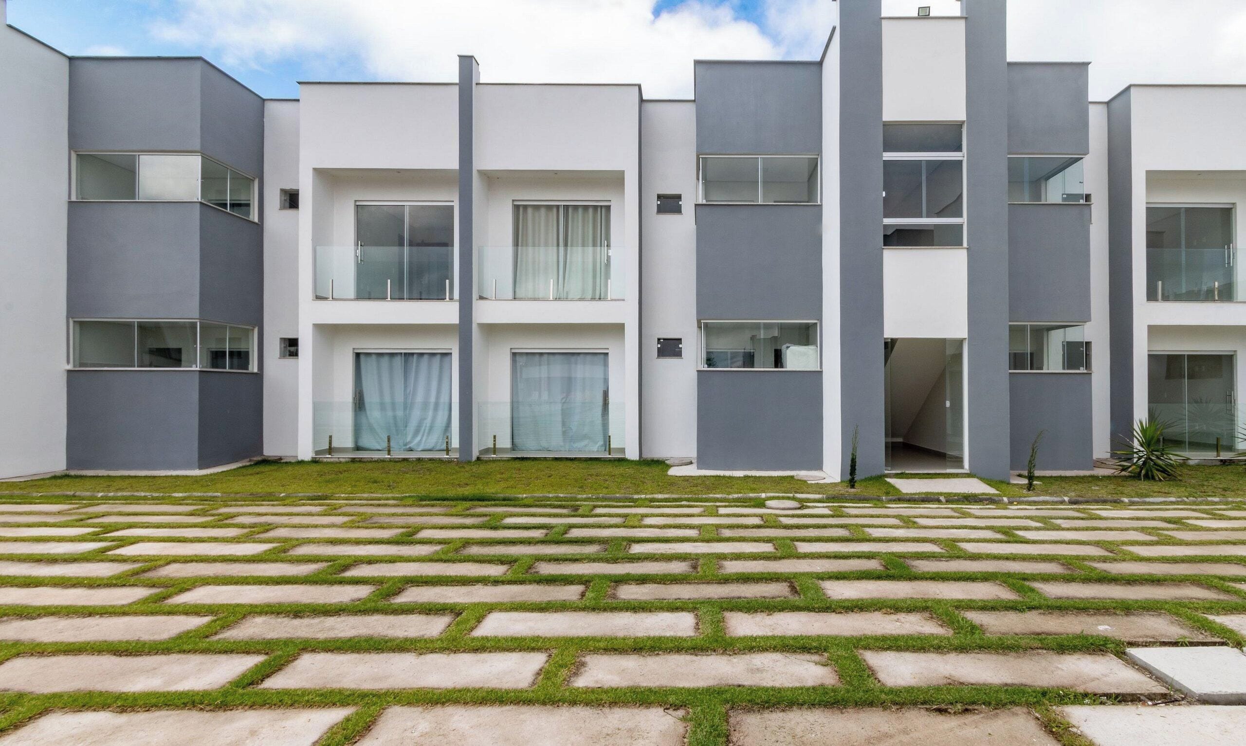 A modern multi-story residential building with a flat roof, large windows, and a paved courtyard with patterned paving.