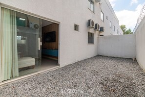 A residential building with a gravel driveway, a sliding glass door leading to a room with a sofa and a television, and air conditioning units on the exterior wall. - Apartments Near Muta Beach, BA - Rfms (Porto Seguro)