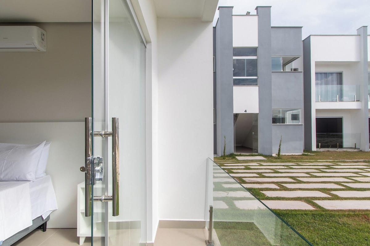 A modern hotel room with a glass door, a bed with white bedding, and a view of a courtyard with a geometric patterned lawn and a building with large windows.