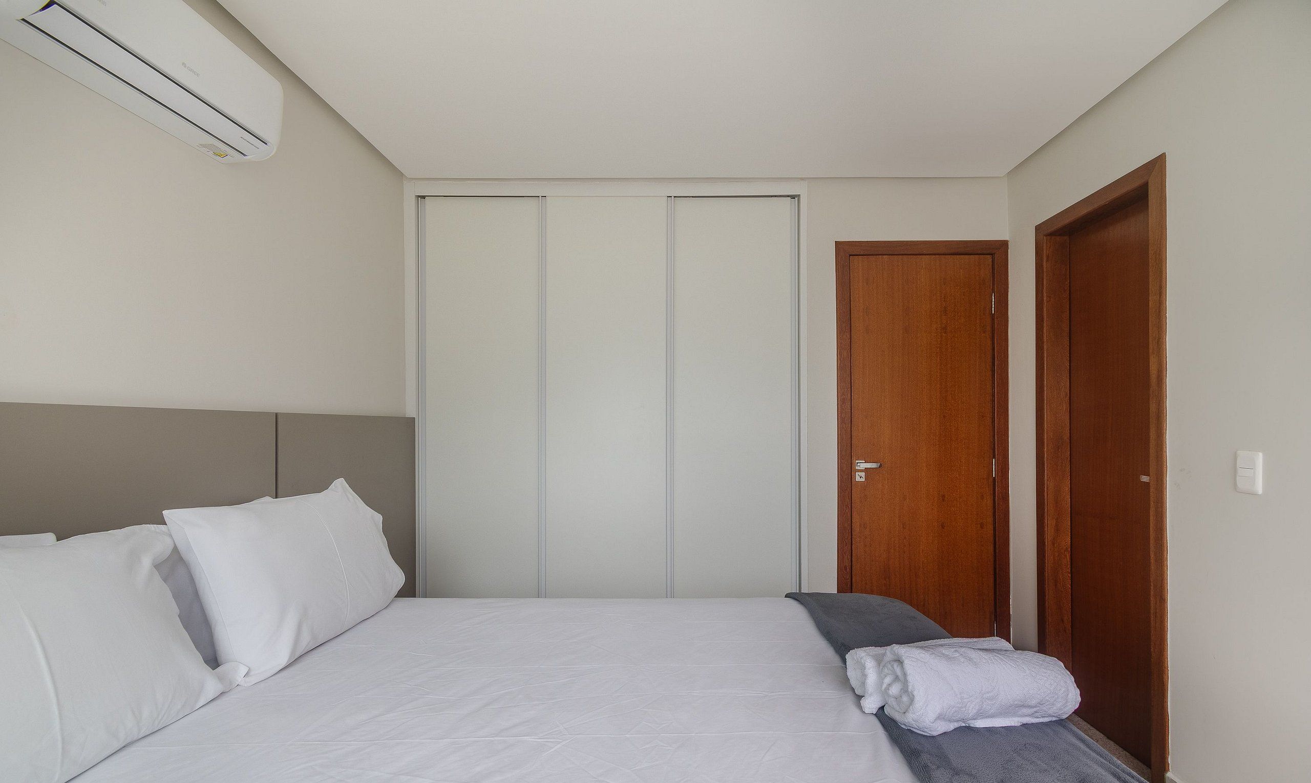 A bedroom with a bed, pillows, a bedside table, a towel, a wooden door, and an air conditioning unit.