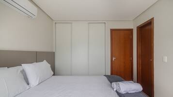 A bedroom with a bed, pillows, a bedside table, a towel, a wooden door, and an air conditioning unit.