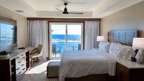 Een hotelkamer met een bed, een bureau, een stoel, een raam met gordijnen, een plafondventilator en uitzicht op de oceaan.
