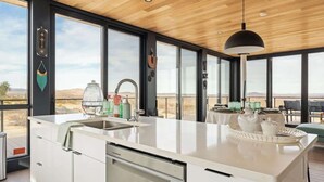 A modern kitchen with a large island, a sink, and a dining area with a view of the desert. - Late for the Sky • Modern Marvel with Hot Tub (Joshua Tree)