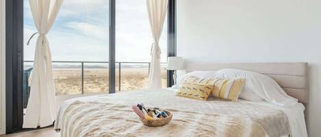 A bedroom with a large bed, a view of the desert through floor-to-ceiling windows, and a basket of toiletries on the bed.