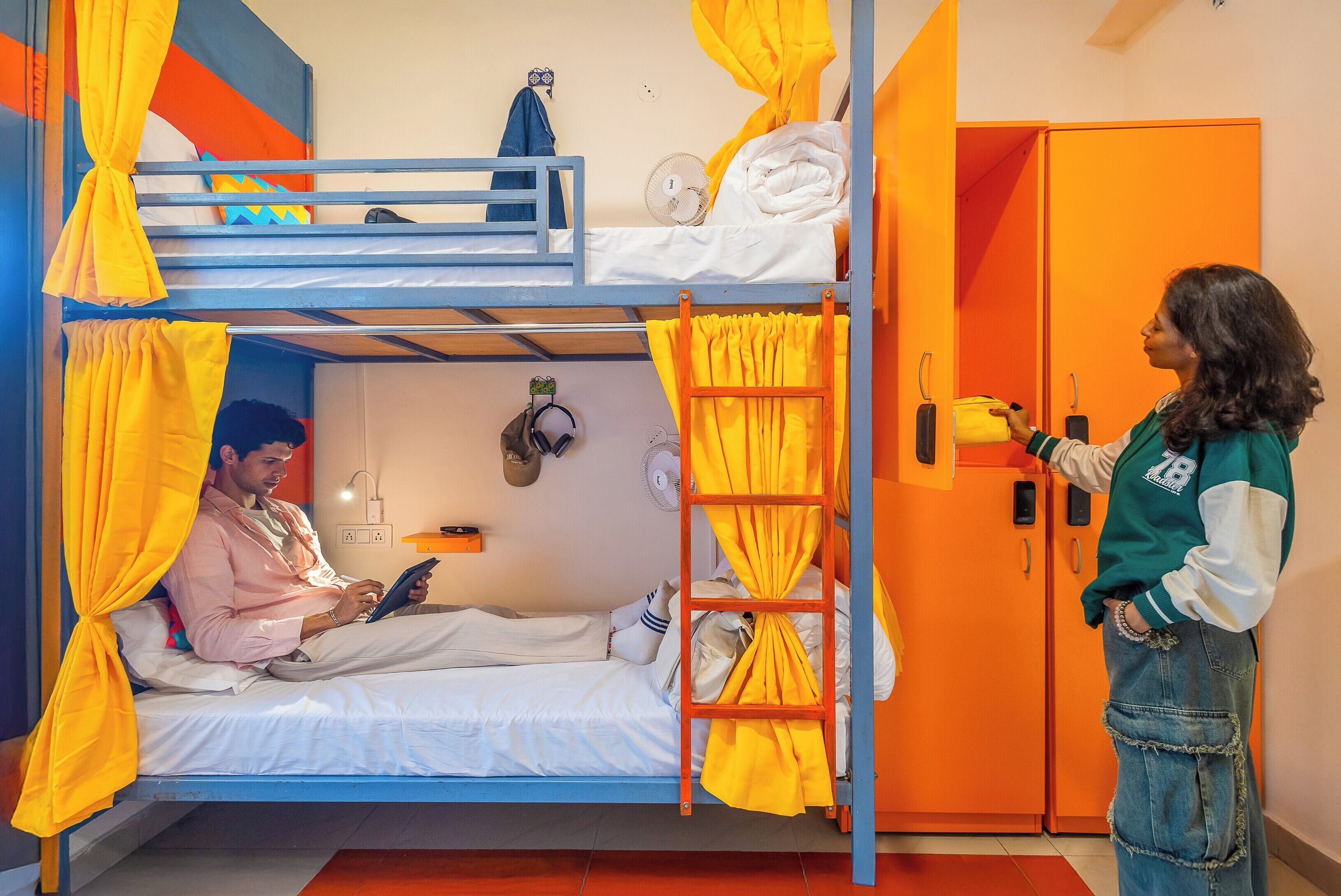A bunk bed room with a person reading a book, another person standing by the door, and a wardrobe.