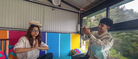Two people sitting on a bed in a room with a view of trees outside. The room has a colorful wall and a fan on the ceiling.