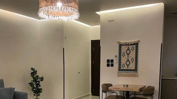 A modern living room with a grey sectional sofa, a dining table with four chairs, a hanging chandelier, and a patterned rug. - Haza Manzeli Rabwa Branch (Madinah)