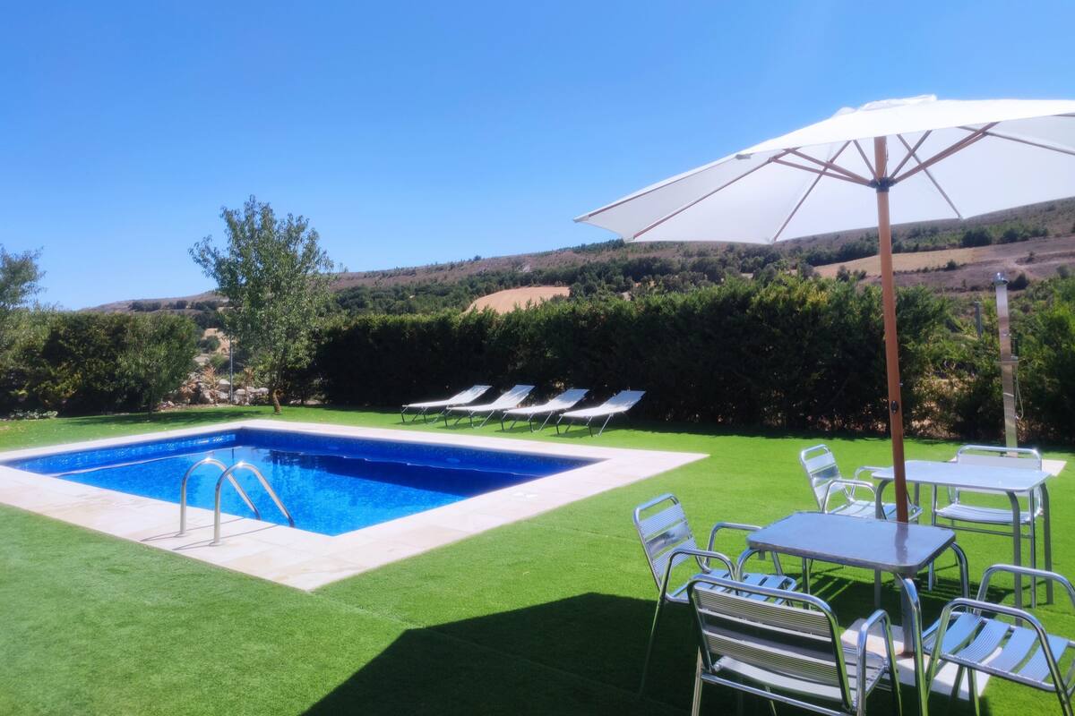 Piscina com vista para colinas, cercada por grama sintética, e uma mesa com cadeiras sob um guarda-sol branco.