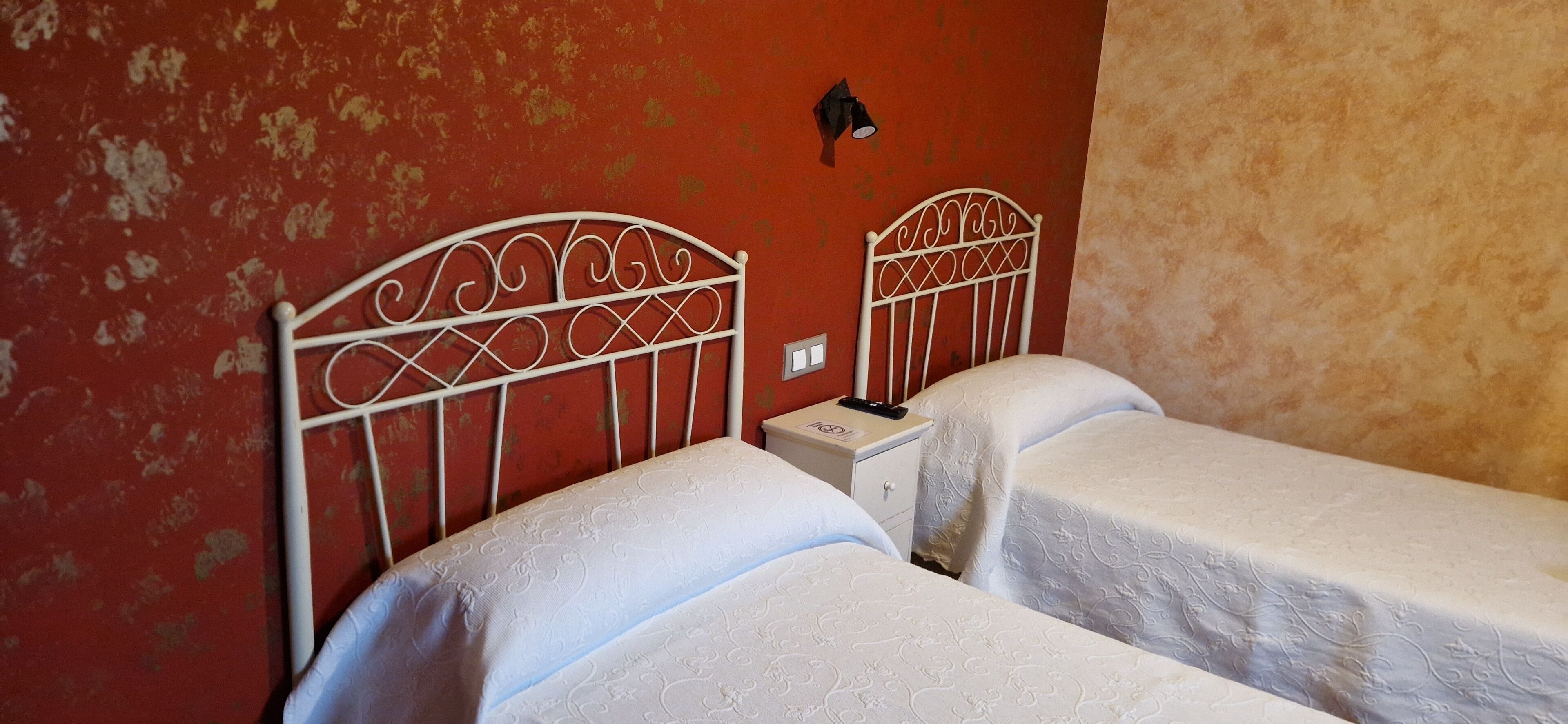 A room with two single beds, a nightstand with a lamp, and a red wall with peeling paint.