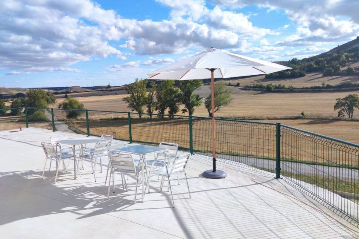 Um pátio externo com mesas e cadeiras, um guarda-sol branco e vista para colinas ondulantes.