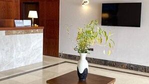 Un espace d’accueil comprenant un comptoir de réception en bois, un vase blanc contenant une plante, une table avec un cadre doré et un téléviseur à écran plat fixé au mur.