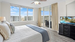 A hotel room with a large bed, a ceiling fan, a TV, and a view of the city through large windows.