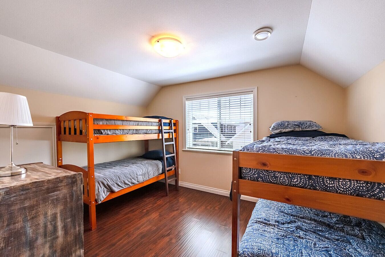 A dormitory room with two bunk beds, a desk with a lamp, a window with blinds, and a ceiling light.