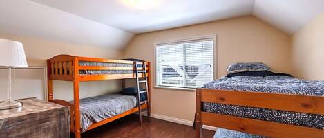 A dormitory room with two bunk beds, a desk with a lamp, a window with blinds, and a ceiling light.