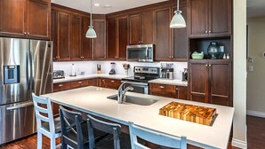 Eine moderne Küche mit Holzausstattung, Edelstahlgeräten, einer zentralen Kochinsel inklusive Barhocker, Pendelleuchten und einem eingebauten Mikrowellenherd.