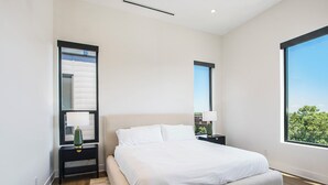 A modern bedroom with a large bed, a nightstand with a lamp, a mirror, and a window with a city view.