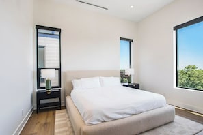 A modern bedroom with a large bed, a nightstand, a lamp, and a window with a view.