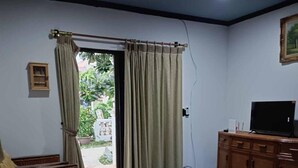 Sala de estar con mobiliario de mimbre, una mesa de centro de vidrio, un televisor sobre un mueble de madera y vista a un jardín por una puerta.