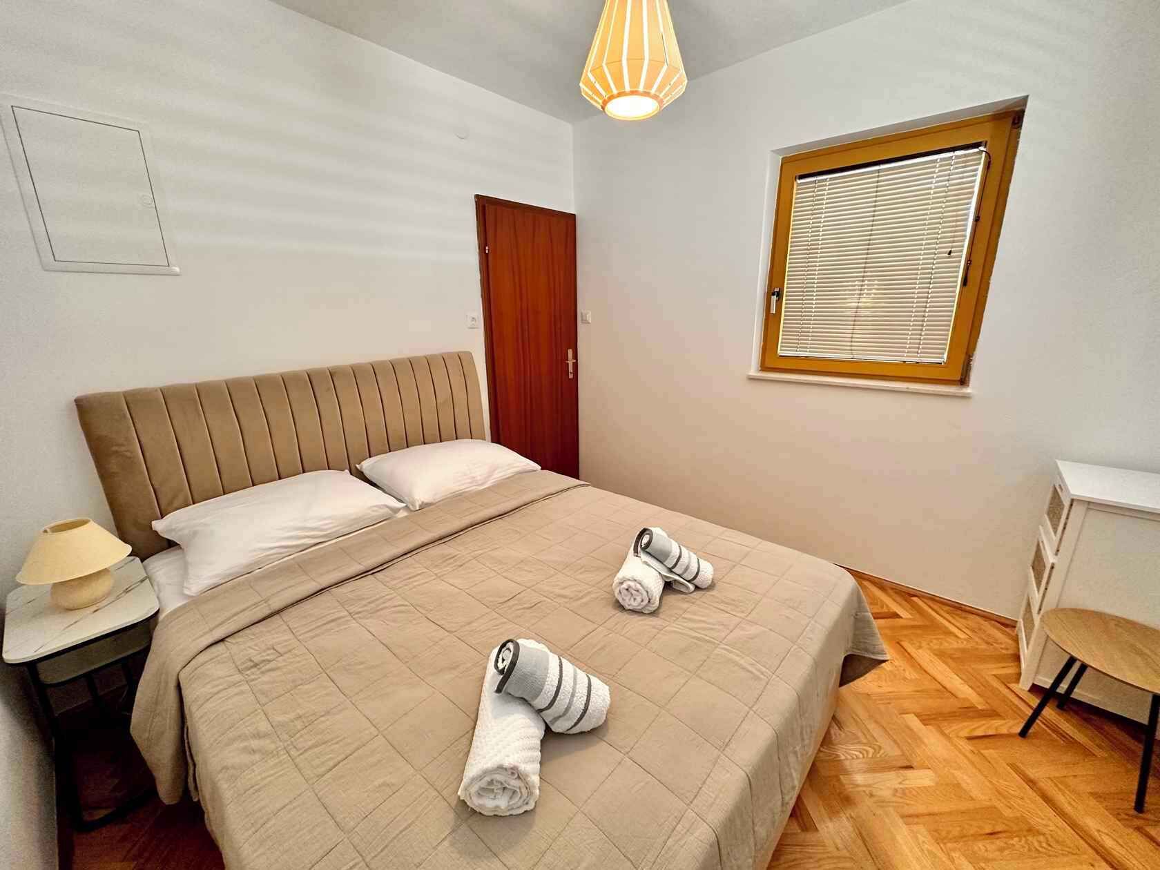 A bedroom with a bed, a nightstand, a desk, a window with blinds, and a hanging light fixture.