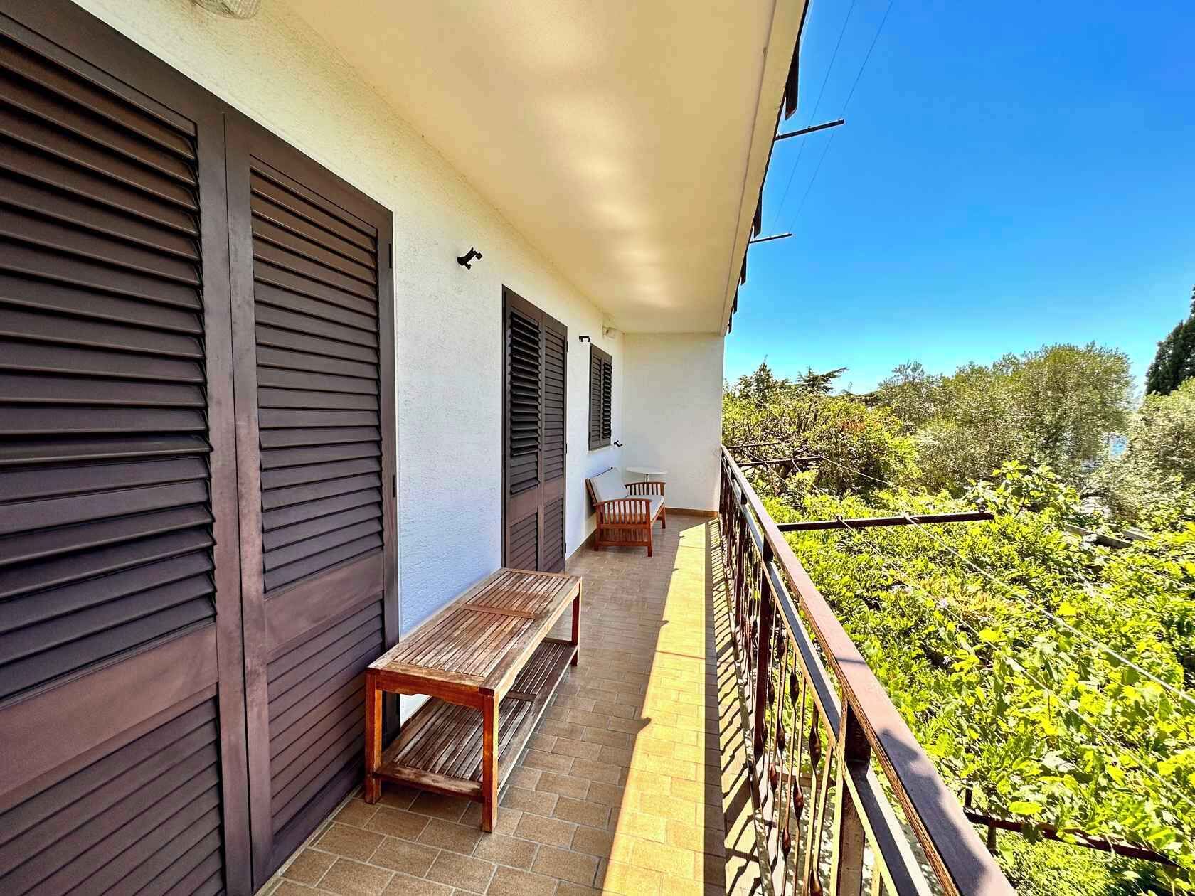 A balcony with a wooden bench and table, a railing, and a view of greenery.