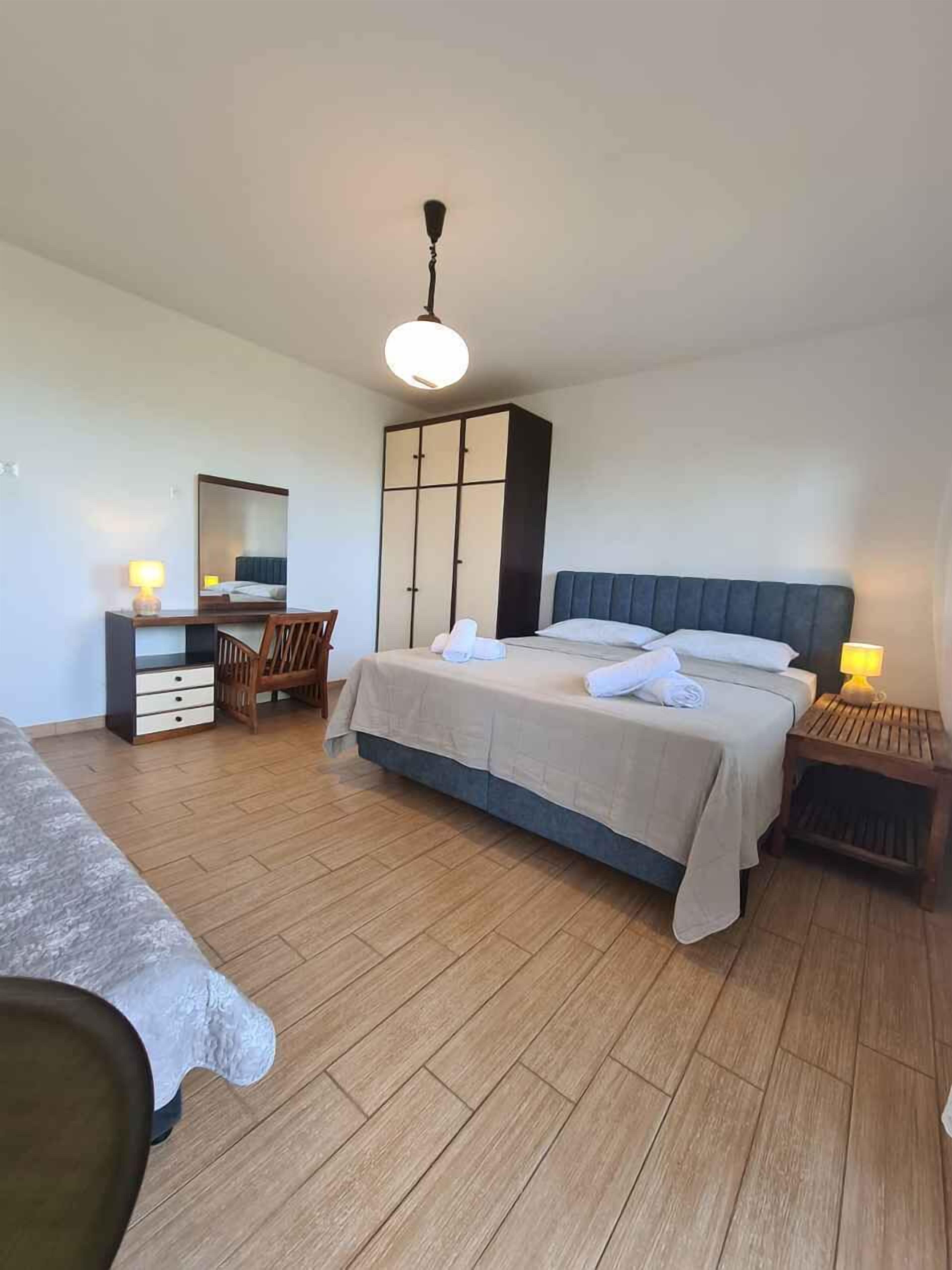 A bedroom with a bed, a desk, a chair, a wardrobe, and a bedside table with a lamp.