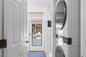Un pasillo con una puerta blanca, vista a un patio trasero a través de una ventana y una lavadora.