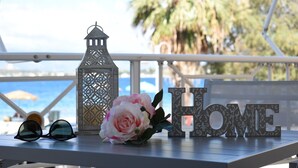 Une table ornée d’une lanterne décorative, d’une fleur, de lunettes de soleil et du mot « MAISON », sur un balcon offrant une vue sur la mer et les palmiers.