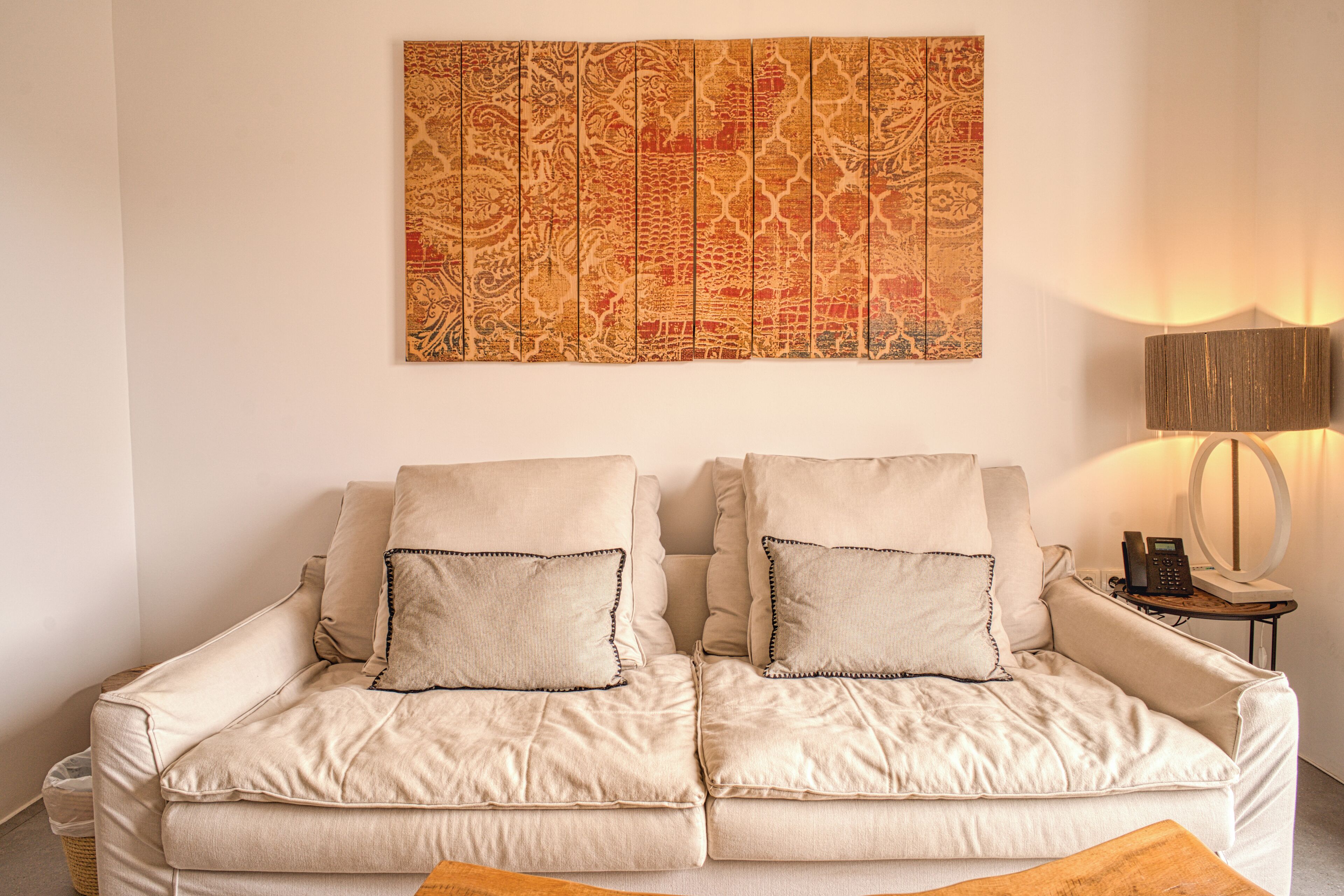 A beige sofa with matching cushions, a decorative tapestry, a lamp, and a small table with a phone.