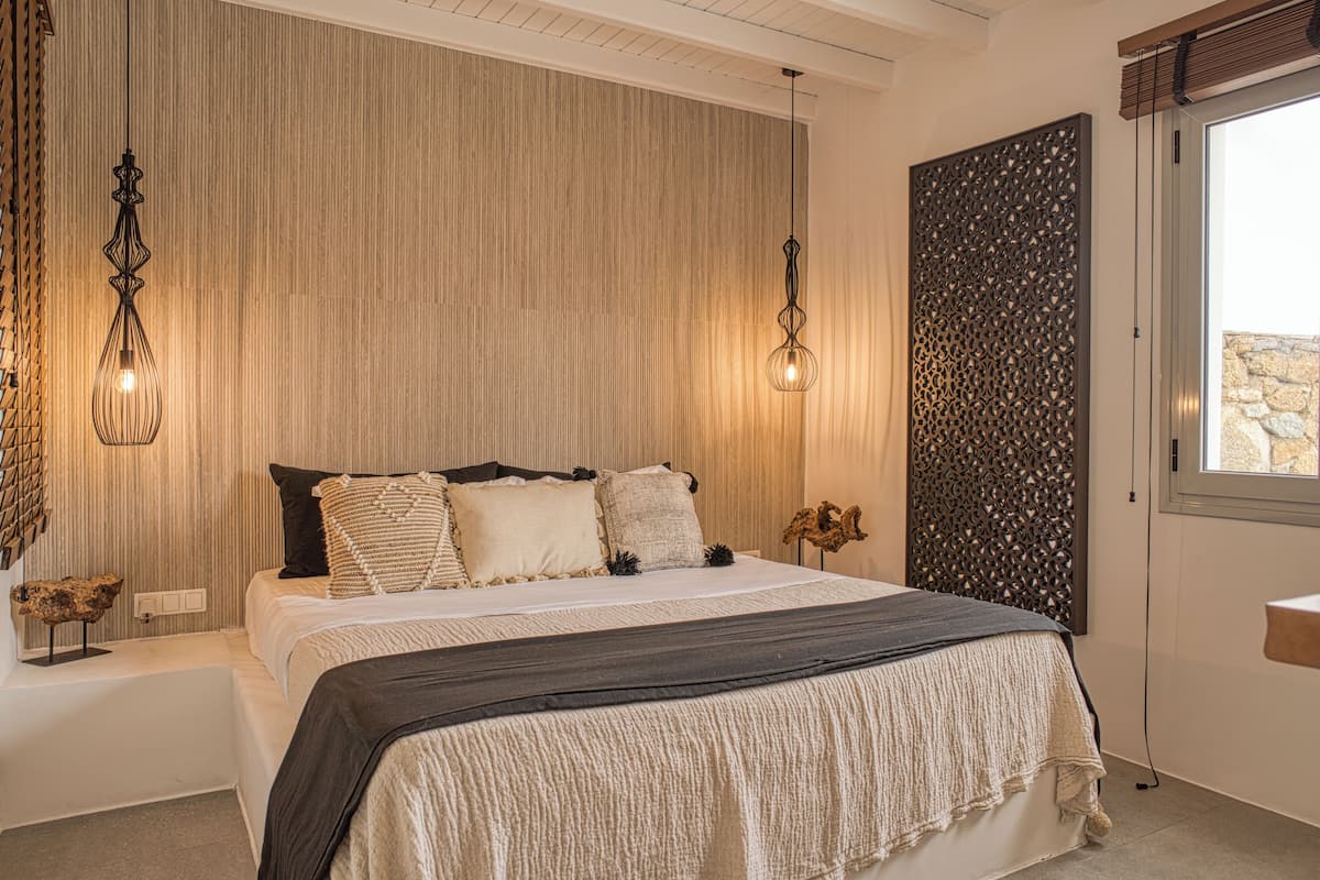 A bedroom with a large bed, two wall-mounted lights, a decorative panel, and a window with a stone wall view.