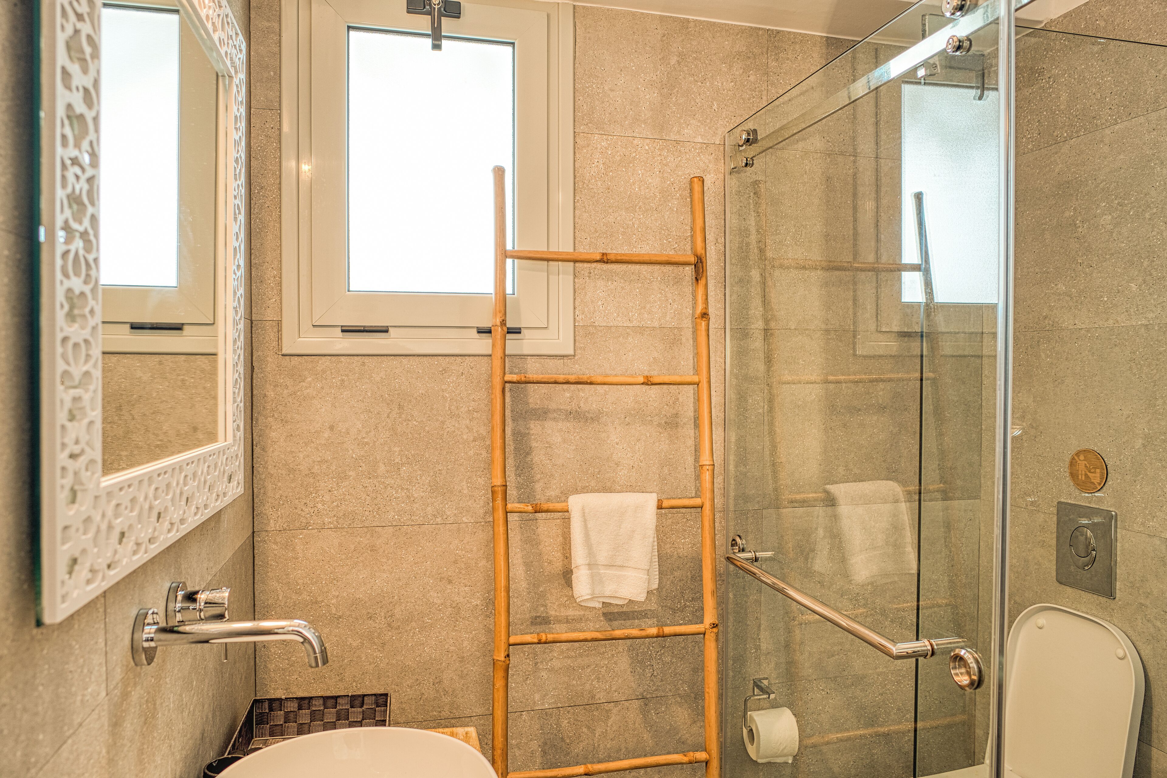 A bathroom with a glass shower, a white sink, a mirror with an ornate frame, and a wooden ladder.