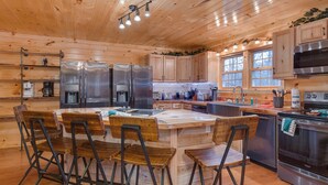 A wooden cabin kitchen with a central island, bar stools, stainless steel appliances, and large windows.